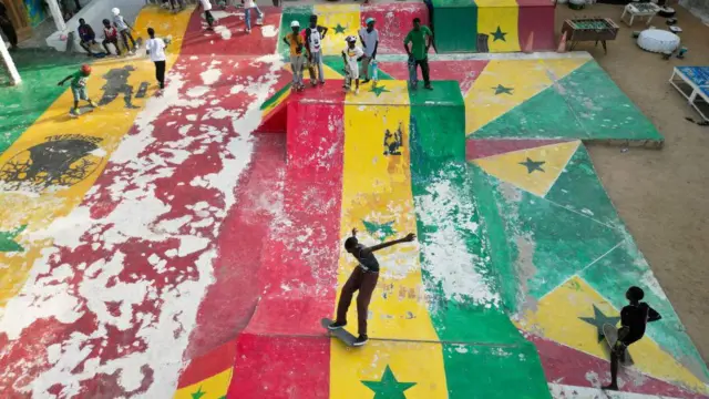 Vue plongeante d'un skatepark animé et coloré du quartier de Ngor à Dakar, au Sénégal. Le parc est un véritable centre névralgique où se côtoient skateurs, tantôt en pleine action sur les rampes, tantôt en train de les observer. L'intégralité du revêtement en béton, y compris les transitions courbes et les rampes abruptes, est peinte aux couleurs vives du drapeau sénégalais : vert, jaune et rouge.