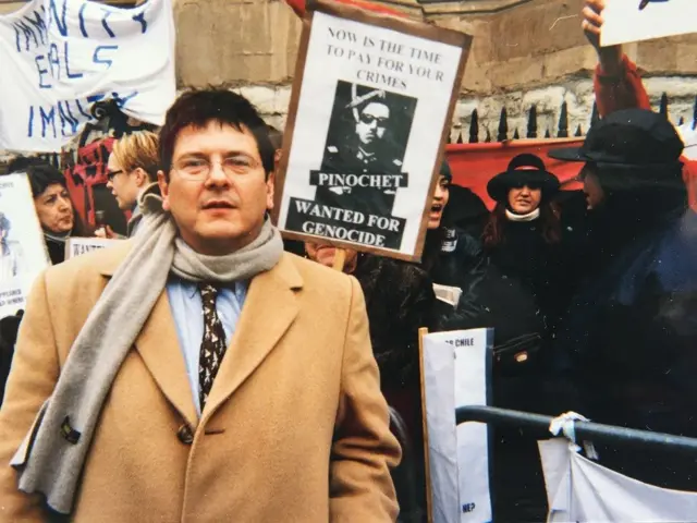 Reed Brody, con un abrigo beige, junto a un grupo de manifestantes, uno de ellos tiene una pancarta con la foto de Pinochet que dice en inglés: "Now is the time to pay for your crimes. Pinochet. Wanted for genocide" ("Llegó el momento de que pagues por tus crímenes. Pinochet. Buscado por genocidio")