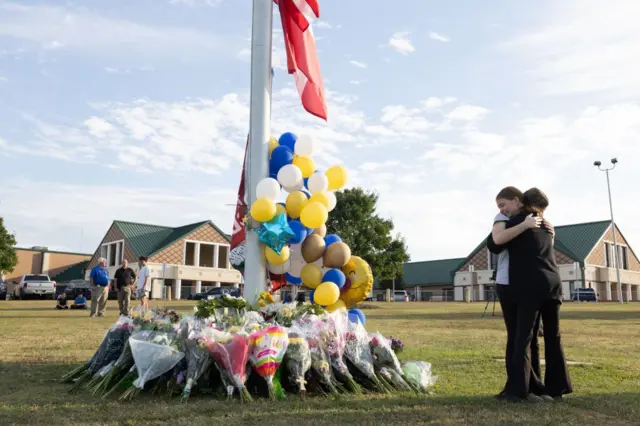 Dos adolescentes se abrazan al lado de un lugar lleno de flores que recuerdan a las víctimas del tiroteo. 