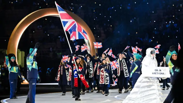 暗い色のそろいの上着と帽子を身に着けた選手たちが英国旗を手にしながらうねった通路を歩いている。それぞれ、クリーム色に青と赤で「GB」、あるいは紺とえんじ色で「GREAT BRITAIN」と大文字で描かれた大きいスカーフを巻いている。