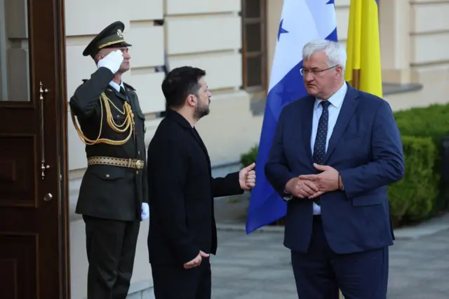 Сибіга у діловому костюмі з краваткою стоїть навпроти Володимира Зеленського у чорному піджаку 
