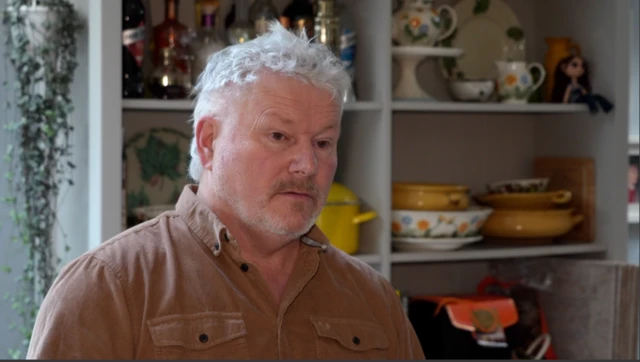 Imagen de un hombre con cabello corto grisáceo y barba. Lleva una camisa marrón con botones negros.