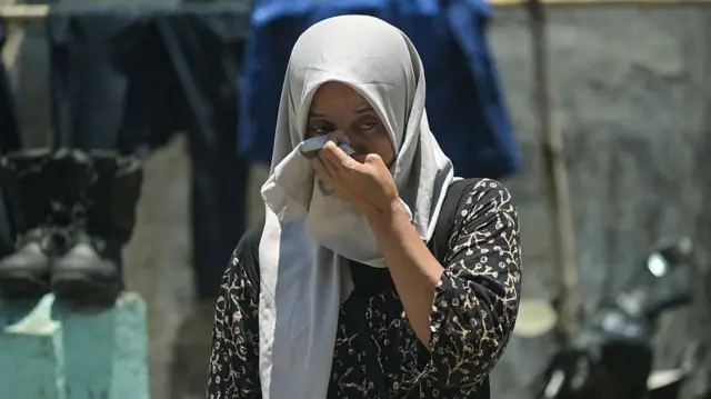 ねずみ色の長い布で頭部を覆った人が、左手で布の一部を鼻の近くに押し当てている