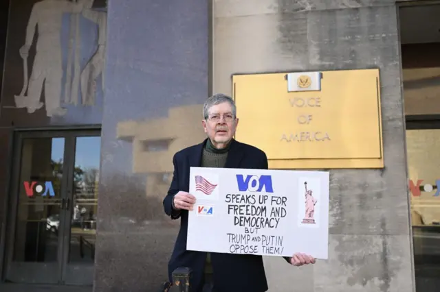 فردی جلوی در ورودی صدای آمریکا در واشنگتن، پایتخت آمریا به بسته شدن صدای آمریکا اعتراض میکند. بر روی پلاکاردی دست این مرد است نوشته شده: صدای آمریکا در حمایت از آزادی و دموکراسی سخن میگوید، اما ترامپ و پوتین با آنها مخالفند