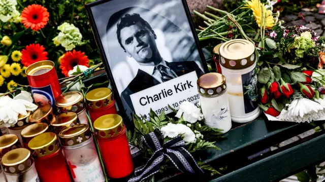 Retrato de Charlie Kirk em preto e branco rodeado de velas e flores. 