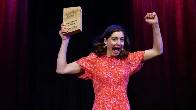 JoJo Maberly, con un vestido rojo, anima mientras sostiene un premio de madera. Tiene cabello castaño y una expresión alegre. El fondo es negro.