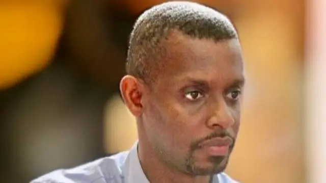 Bukuru Muhizi, vêtu d'une chemise bleu clair, détourne le regard de la caméra.