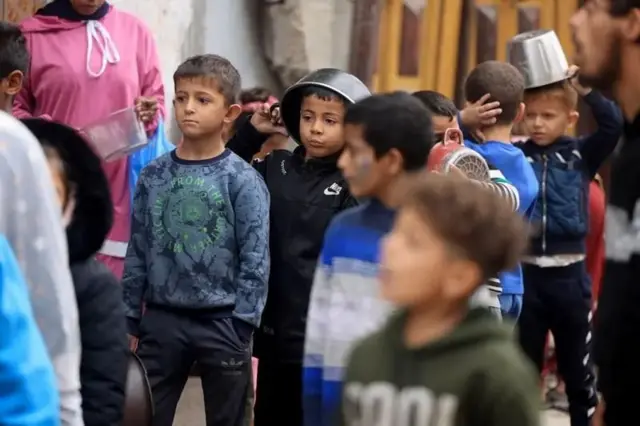 صفی از کودکان که برای غذا قابلمه در دست دارند