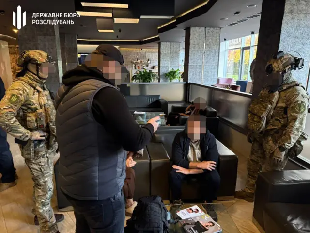 Затриманий чоловік у кріслі в лоббі готелю, його обличчя заблюрене. Поруч – двоє чоловіків у військовій формі та повному екіпіруванні, на передньому плані – чоловік у чорному цивільному одязі спілкується з затриманим.