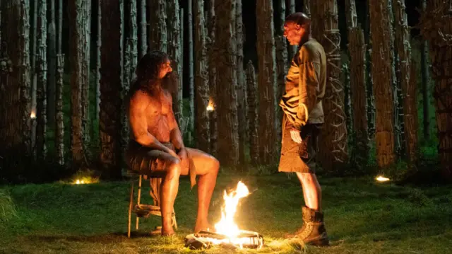 Ральф Файнс и другой персонаж в лесу по обе стороны от небольшого костра