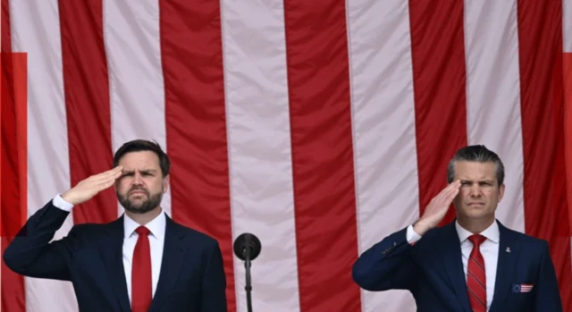 JD Vance ve Pete Hegseth  Amerikan bayrağı önünde asker selamı veriyor. Üzerlerinde takım elbise var.