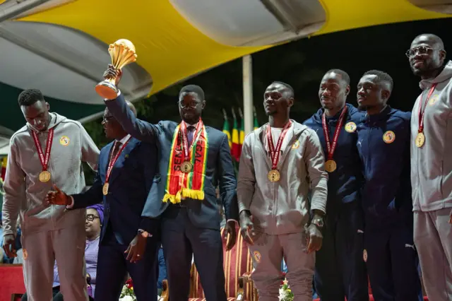 Le président Bassirou Diomaye Faye soulève le trophée de la CAN 2025 avec sa main droite, avec autour de lui des joueurs de la sélection et des membres du staff.