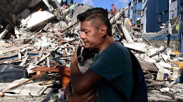 Un hombre abraza a una mujer y detrás los escombros de un edificio derruido.