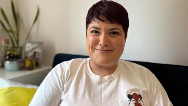 У Ді коротке темне волосся, вона сидить на темному дивані, одягнена в білу футболку з логотипом «Справедливе ставлення до жінок Уельсу» праворуч. На логотипі зображено трьох жінок-палиць, які з'єдналися за руки та тримають рівновагу, немов терези. На задньому плані ліворуч зображені рослини та прикраси на полиці.