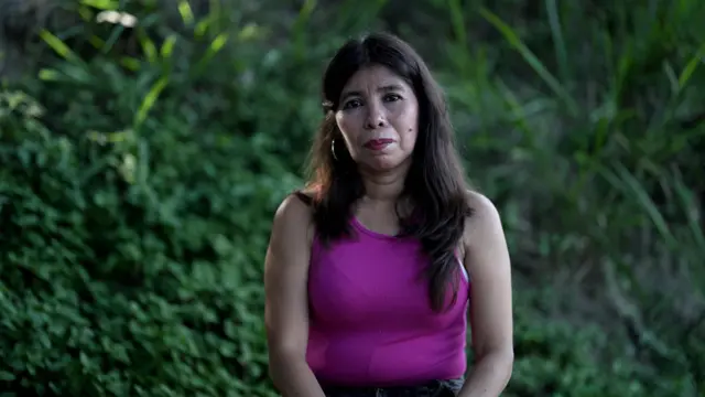 Mulher com blusa rosa com plantas ao fundo.