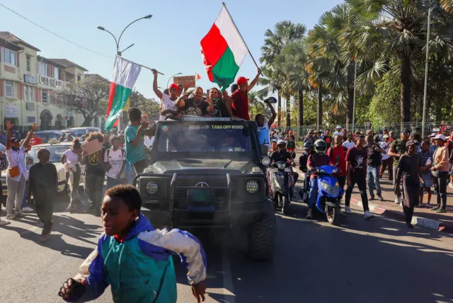 صبي يركض أمام شاحنة صغيرة مع أشخاص يلوحون بالأعلام بعد انضمام الجيش إلى المتظاهرين المتجمعين خارج مبنى البلدية في شارع الاستقلال خلال مظاهرة وطنية بقيادة الشباب ضد انقطاع التيار الكهربائي المتكرر ونقص المياه، في أنتاناناريفو، مدغشقر، 14 أكتوبر/تشرين الأول 2025.