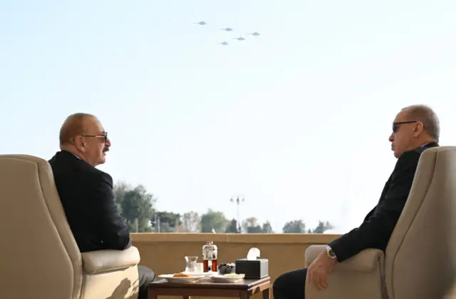 Вид со спины. Президенты Азербайджана и Турции сидят в креслах на фоне самолетов в небе.