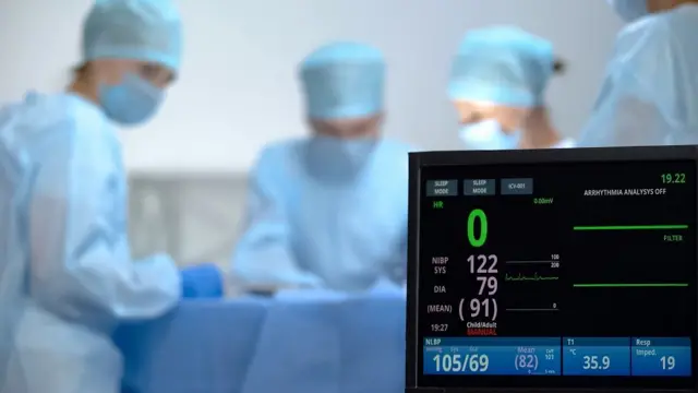 Médicos em uma sala de cirurgia com um monitor que não mostra frequência cardíaca.