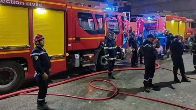 Des pompiers en uniforme bleu se tiennent devant des camions de pompiers.