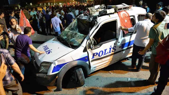 15 Temmuz Ankara'da sokağa çıkan kalabalıklar. Onlarca kişi, parçalanmış bir polis aracının etrafında görülüyor. Bazılarının elinde Türk bayrakları var. Hepsi erkek. 