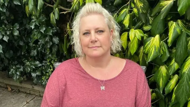 Clare is sitting on a chair, looking straight at the camera. She has short, blonde, curly hair. She is wearing a dark pink, short-sleeved T-shirt and a silver necklace. She is outside and there are bushes in the background.