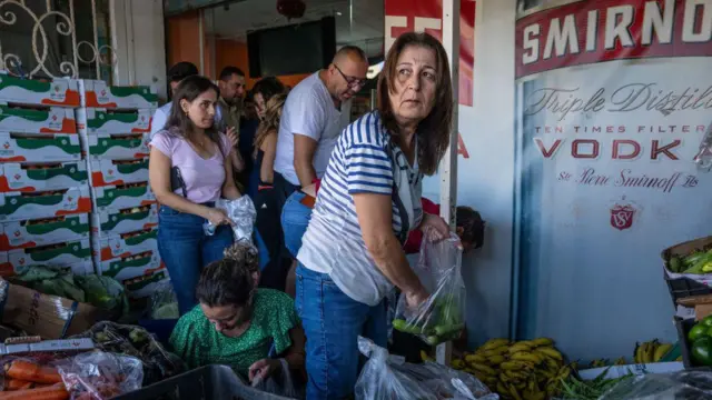 أشخاص يصطفّون داخل متجر صغير للخضار، يختارون منتجاتهم من صناديق مكدّسة؛ امرأة ترتدي قميصاً مخططاً تضع فلفلاً أخضر في كيس في المقدّمة، فيما ينتظر آخرون خلفها. موز وخضروات متنوّعة عند المدخل، ,ويغطي إعلان لفودكا جزءاً من الجدار.