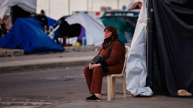 سيدة محجبة ترتدي ملابس باللون الأحمر تجلس على كرسي أمام خيمة باللون الأبيض، وخلفها خيام نازحين أخرى