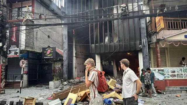 पोखरा लेकसाइडमा आगजनी गरिएको एक होटल र त्यसको अगाडि दुई विदेशी पर्यटक