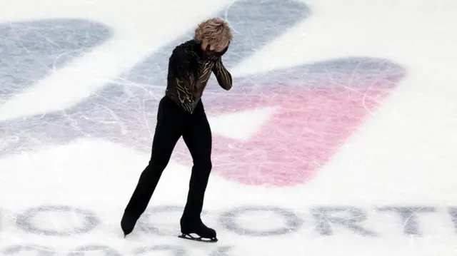 黒い衣装のマリニンが顔を両手で覆いながらリンクを滑っている