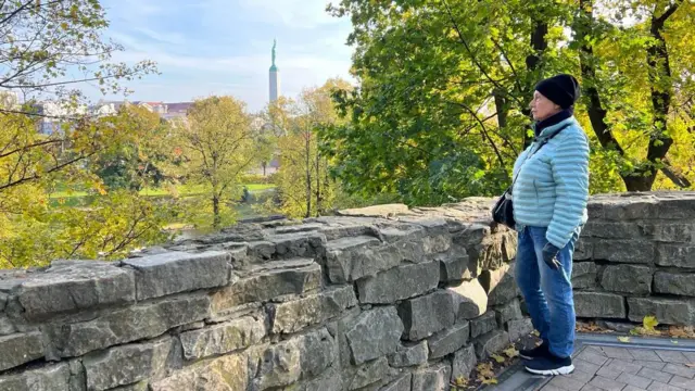 Пожилая женщина в голубой куртке, черной шапке и джинсах стоит в рижском парке. Вдали виден памятник Свободы