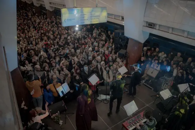 Великий натовп людей, зібраних на станції метро. У центрі – невелика сцена з музикантами. Біля платформи стоїть електропоїзд. Над натовпом висить інформаційний знак із назвами станцій. Освітлення яскраве, видно прожектори та апаратуру для виступу. 