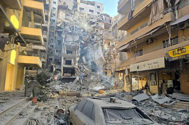 مبنى مدمر وسط مبانٍ أخرى، حيث تظهر آثار الدمار في الشارع، ويظهر في الصورة الملتقطة نهاراً تضرر إحدى السيارات.