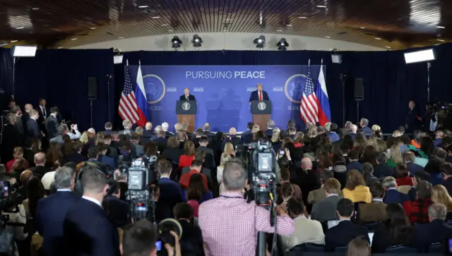 Los periodistas se encuentran frente a un escenario con Putin y Trump de pie en podios. Dos cámaras sobre trípodes están en primer plano, operadas por dos hombres. Los demás periodistas están sentados, muy juntos.