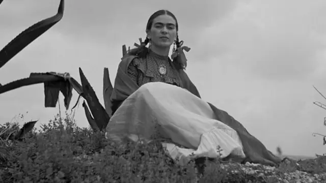 Foto em preto e branco de Frida Kahlo, usando tranças no cabelo e sentada sobre a relva