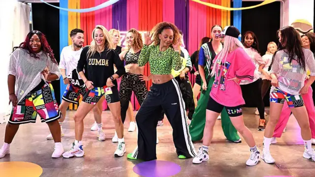 Um grupo de pessoas participa de uma aula de Zumba em um estúdio, dançando em sincronia diante de um cenário colorido. Os participantes usam roupas esportivas vibrantes e executam a coreografia enquanto a instrutora, ao centro, lidera os movimentos.