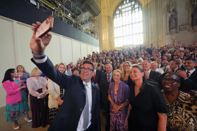 英議会議事堂のゴシック様式の壁や彫像や窓を背に、階段に並ぶ数十人の前で、スマートフォンで自撮りする笑顔のバーナム氏