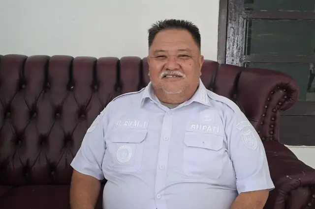 Bupati Bengkulu Selatan, Rifai Tajudi menggunakan baju putih, dan melihat tersenyum ke arah kamera.