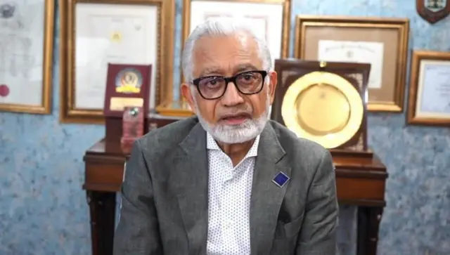 प्रोफ़ेसर डॉक्टर जावेद अकरम कैमरे की तरफ़ देखते हुए, उनके पीछे कुछ शील्ड और फ़्रेम किए हुए सर्टिफ़िकेट नज़र आ रहे हैं.