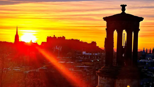 Una vibrante puesta de sol sobre Edimburgo, con el Monumento a Dugald Stewart recortado en primer plano y el Castillo de Edimburgo visible en la distancia.