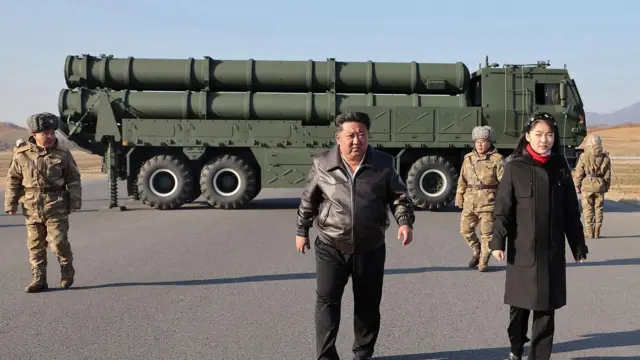 黒い服を着た親子がアスファルト舗装の上を少し離れて横並びに歩いている。背後にはミサイルを積んだ大型車両がある。軍関係者らしき人も数人いる