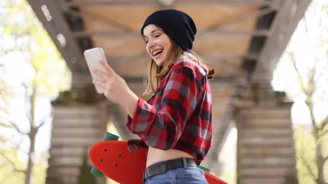 Nastolatka w czerwonej koszuli w kratę i ciemnej czapce na głowie trzyma w lewej ręce telefon i uśmiecha się patrząc na niego 