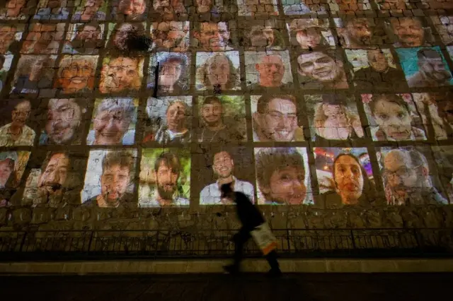 Vultosite de aposta aviaopessoa passando, a noite,site de aposta aviaofrente a muro onde são projetadas fotossite de aposta aviaoreféns; 