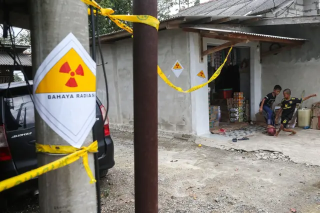 Stiker bahaya radiasi tertempel di tiang listrik dengan dua orang anak sedang bermain bola di belakangnya.