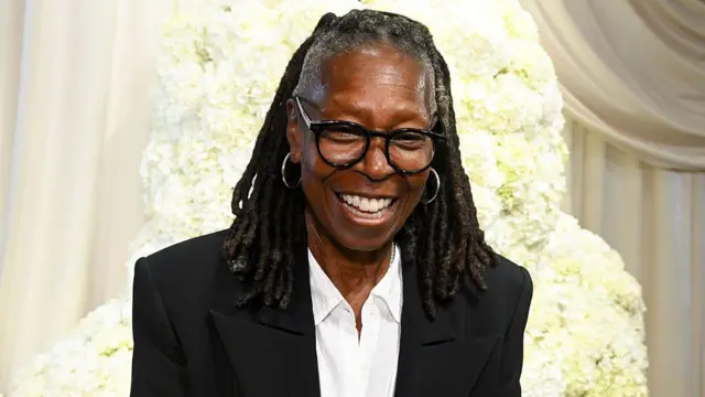 Вупі Голдберг у чорному піджаку та білій сорочці, вона сміється