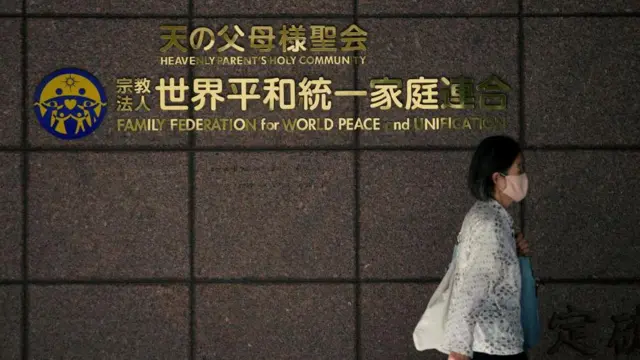 世界平和統一家庭連合(旧統一教会)の文字が張られた壁の前を、マスクをした白い服の女性が歩いている