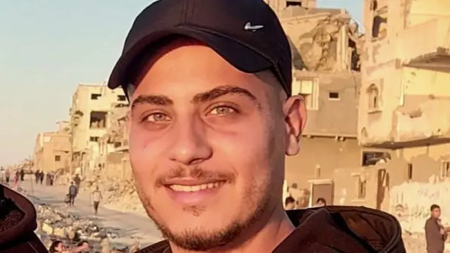 A young man wearing a dark hoodie and a black baseball cap smiles at the camera. In the background is the ruins of damaged buildings