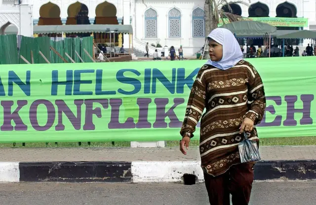 Seorang wanita berjalan di samping spanduk mengenai kesepakatan damai antara pemerintah Indonesia dan kelompok pemberontak separatis Aceh (GAM) yang dipasang di sepanjang jalan di Banda Aceh, 15 Agustus 2005. Pada hari Minggu, para pemimpin pemberontak Aceh menuduh pemerintah Indonesia gagal melucuti senjata milisi di provinsi tersebut—sehari sebelum kedua belah pihak dijadwalkan menandatangani kesepakatan damai bersejarah di Helsinki, yang telah memunculkan harapan akan berakhirnya konflik berdarah selama puluhan tahun di provinsi yang luluh lantak akibat tsunami itu. AFP PHOTO/Bay ISMOYO (Kredit foto harus mencantumkan