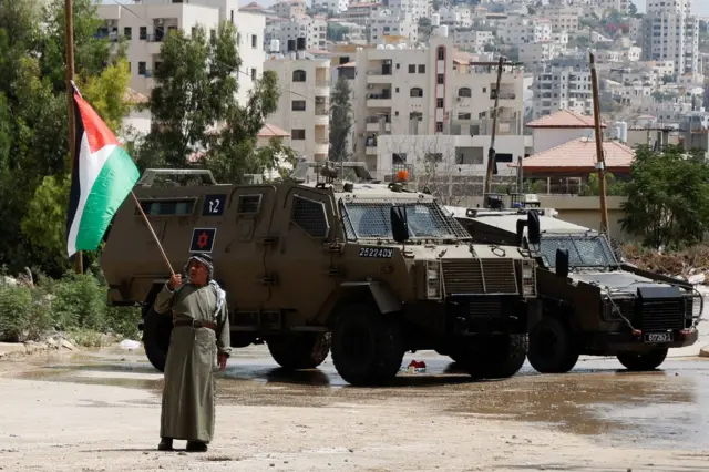 شخص يحمل العلم الفلسطيني أمام مدرعات الجيش الإسرائيلي في مدينة طولكرم في الضفة الغربية.