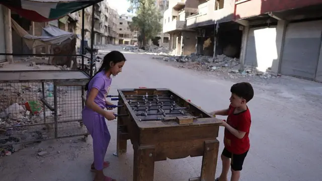 Дети играют в настольный футбол на фоне разрушенных домов и пустых улиц в лагере Ярмук недалеко от Дамаска