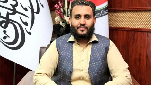 
دکتر شرافت زمان عمار در مطب خود نشسته است. پرچم طالبان در پس زمینه دیده می شود. او پیراهن عاج کم رنگ و کلاهی به تن دارد.
